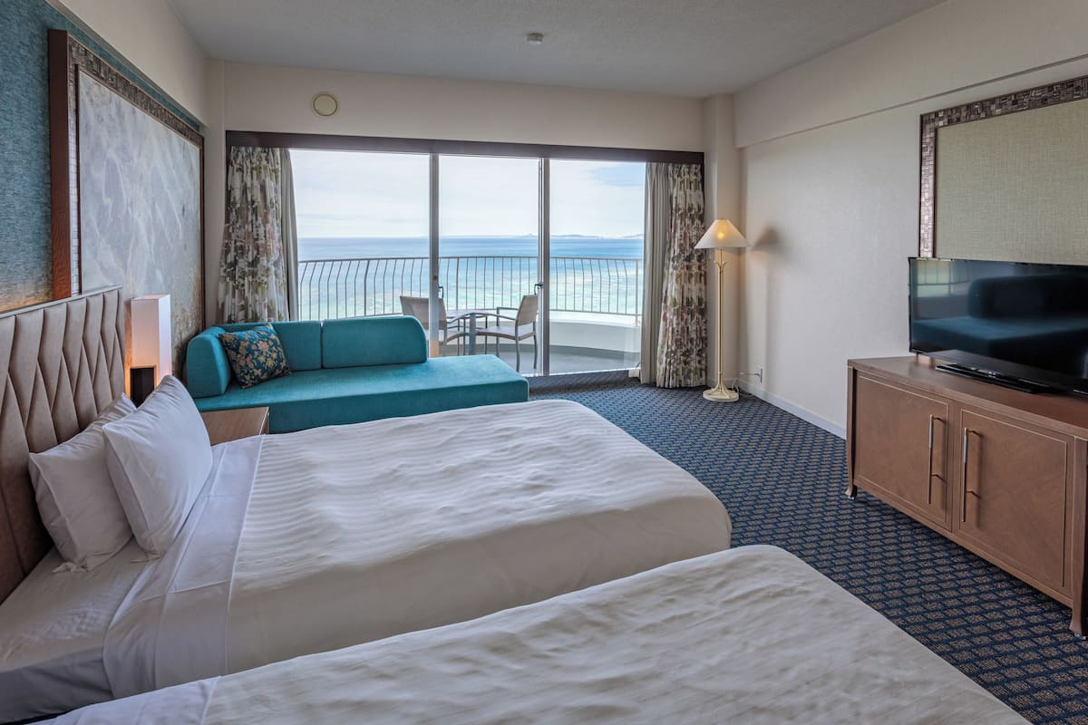 Habitación de hotel con dos camas, un sofá, una televisión y un balcón con vista al mar.