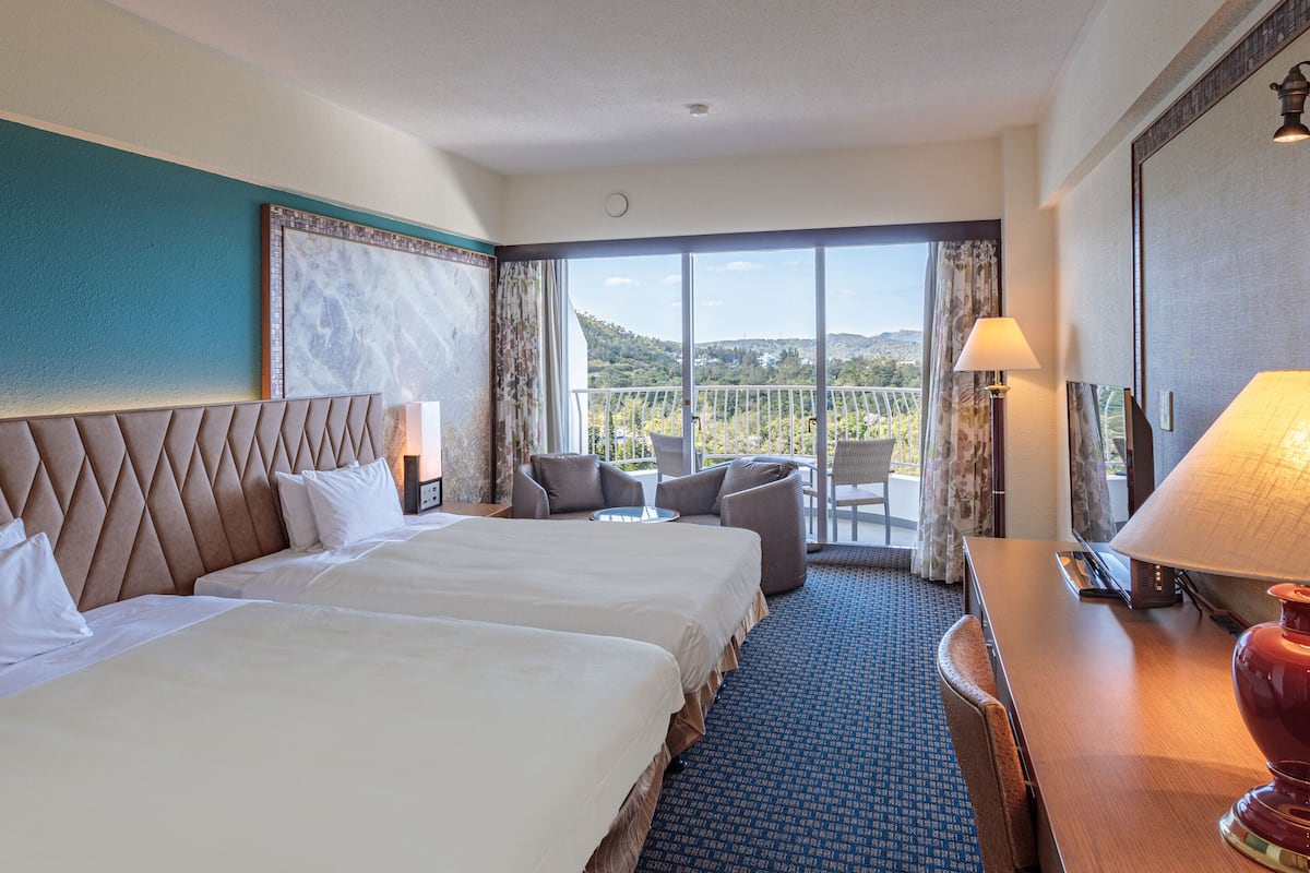 Habitación de hotel con dos camas, un escritorio, una silla y un balcón con vistas.