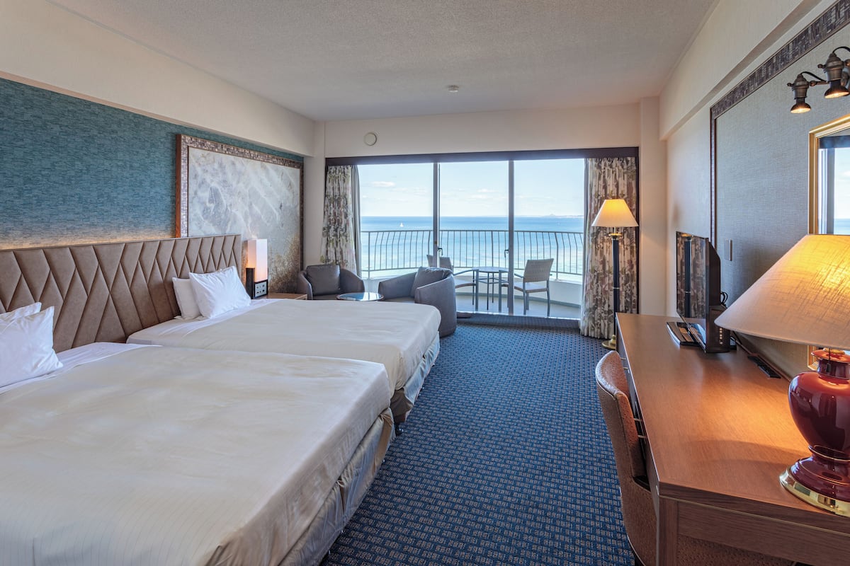 Habitación de hotel con dos camas, un escritorio, una silla, un balcón con vistas al mar y un espejo grande.