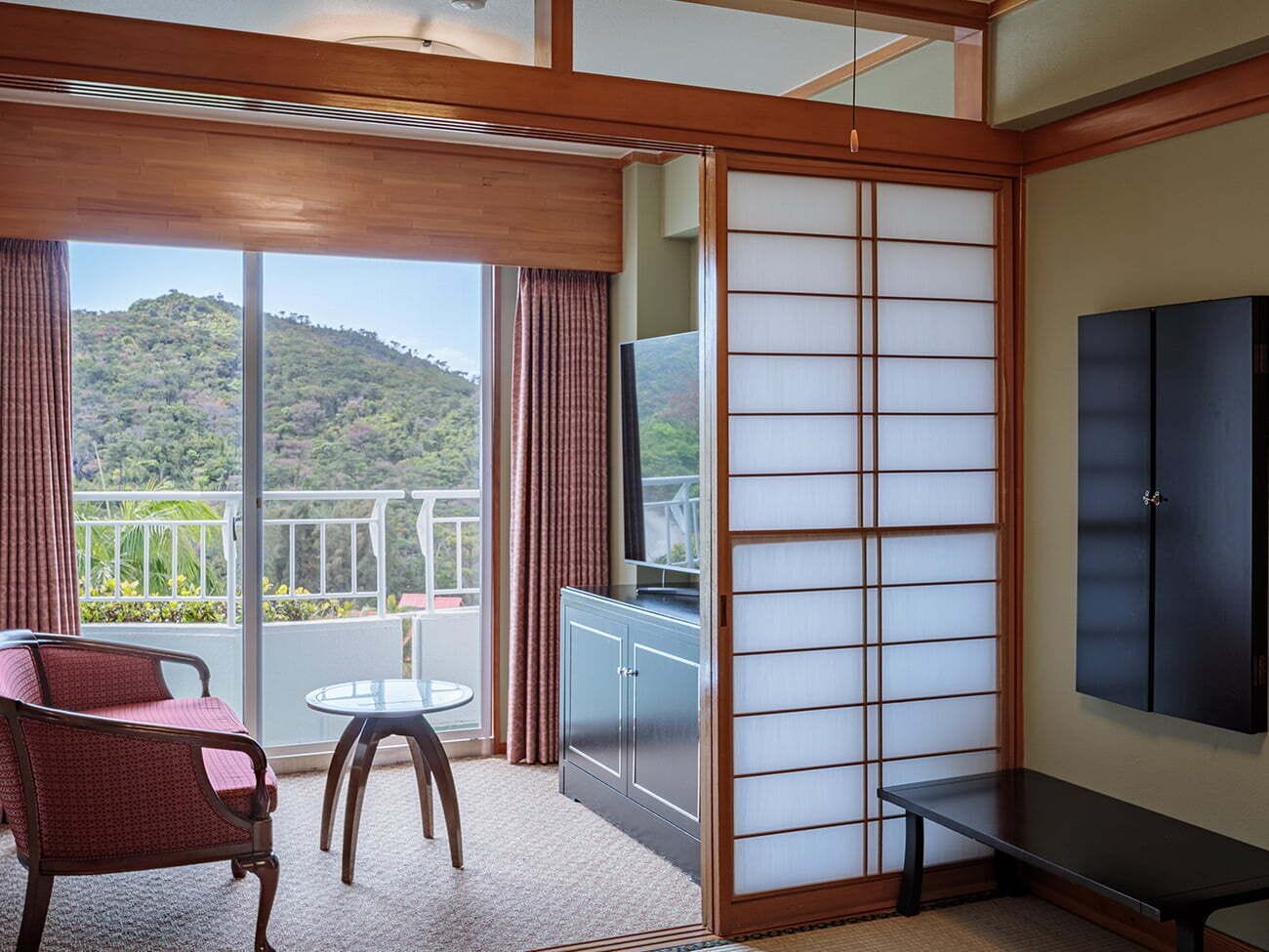A room with a sliding shoji screen, a TV, a sofa, a small table, and a view of a mountainous landscape.