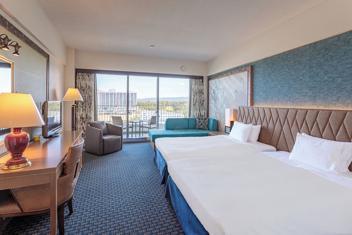 Habitación de hotel con dos camas, un escritorio con lámpara, una silla y vistas a la ciudad a través de amplios ventanales.
