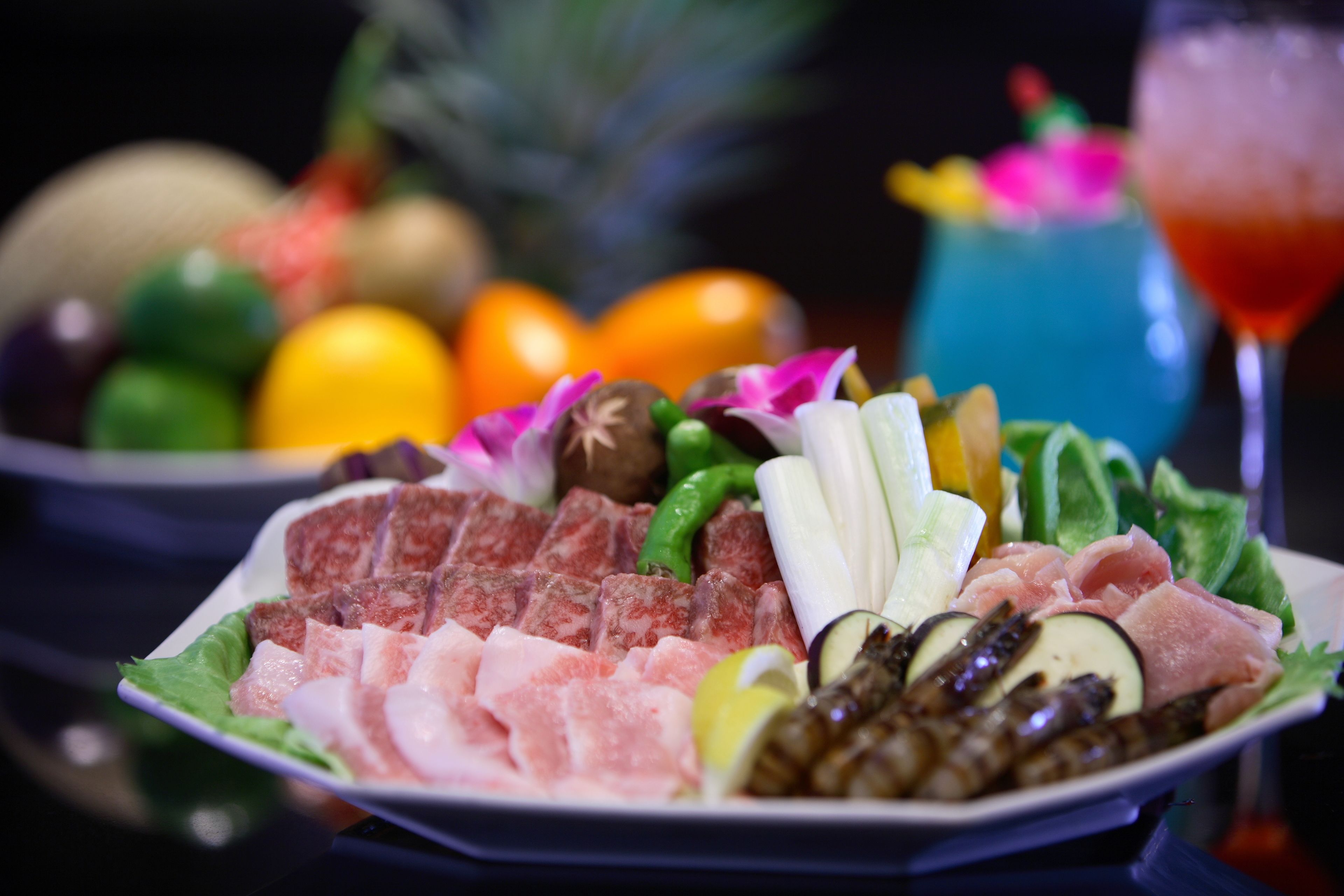 A plate of assorted meats and vegetables, a cocktail glass, and a fruit bowl in the background.