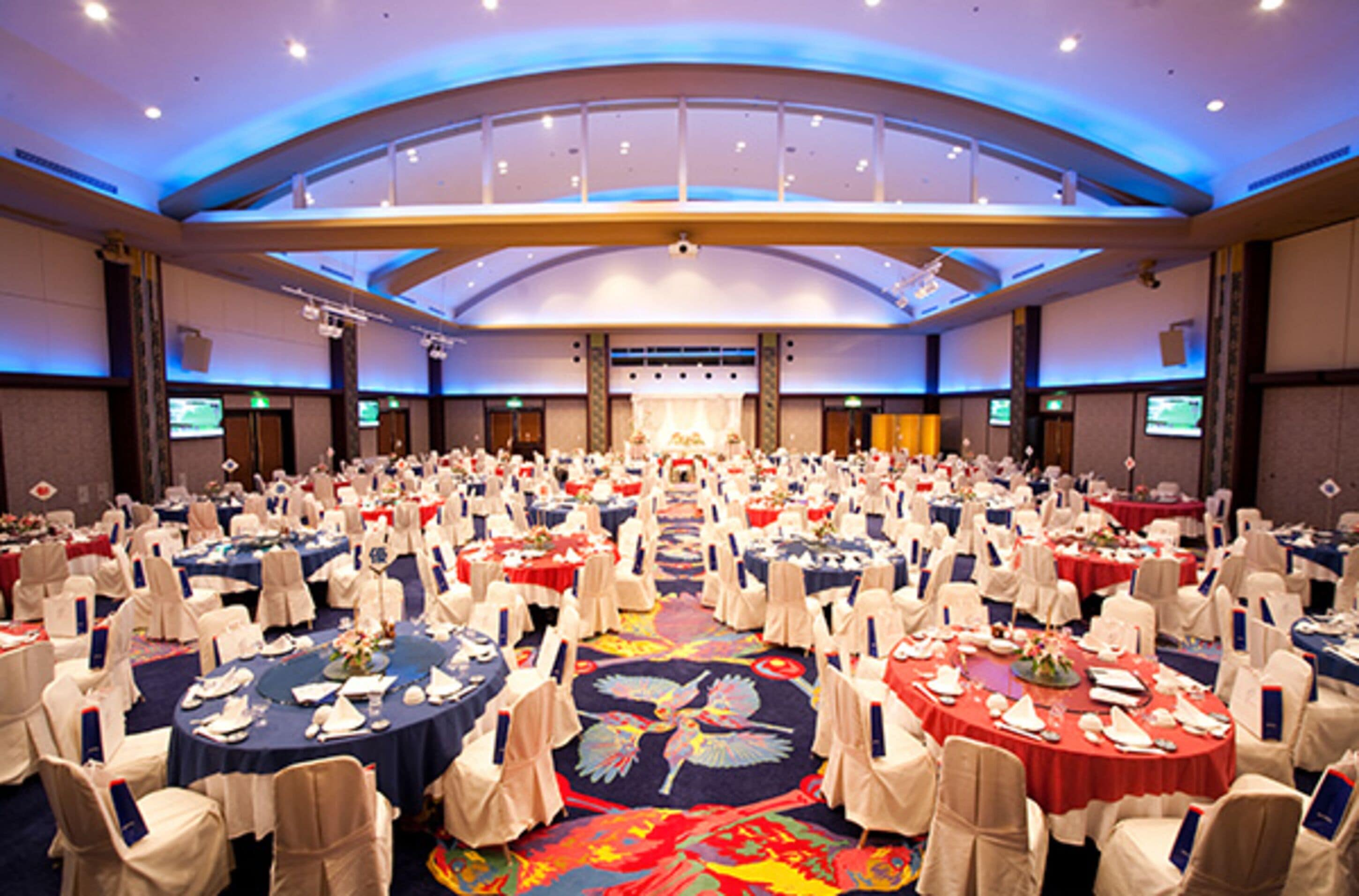 A large conference room with multiple round tables, each set with white tablecloths and chairs, arranged in rows.