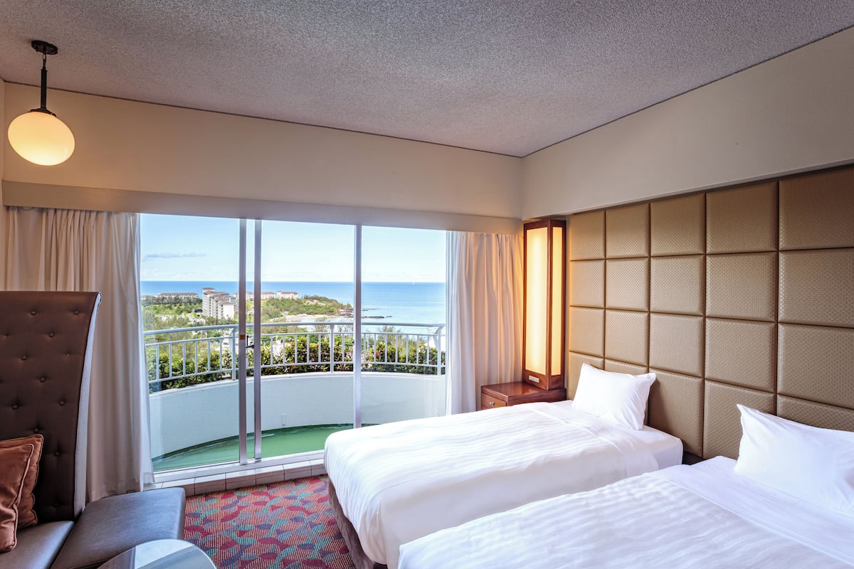 Habitación de hotel con dos camas, un ventanal con vistas al mar, un balcón y una zona de estar también con vistas.