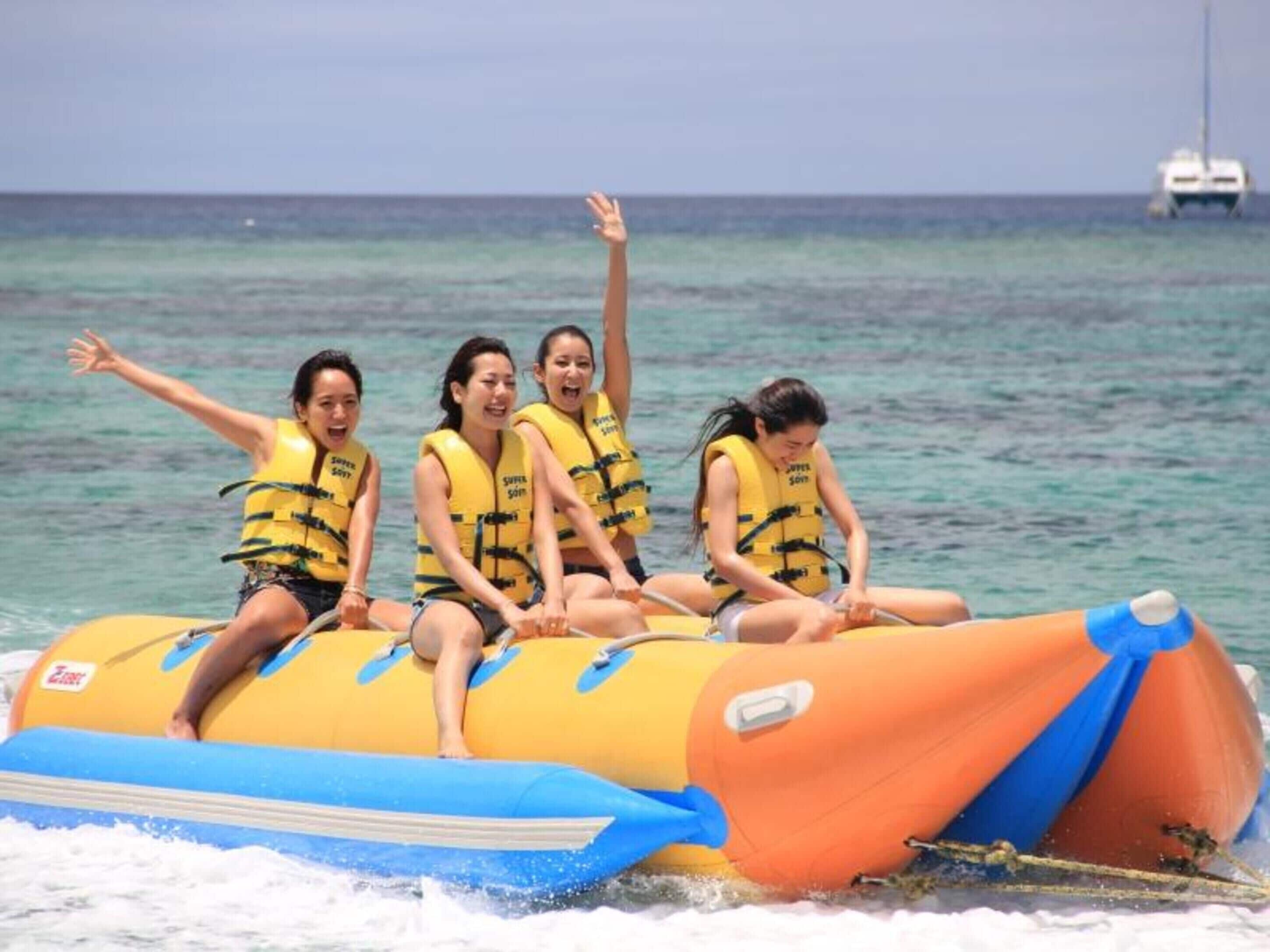 Four people on a yellow and orange inflatable raft with blue trim, wearing yellow life jackets, on a body of water with a ship in the background.
