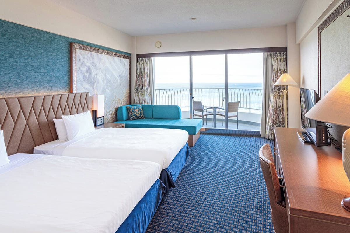 Habitación de hotel con dos camas, un sofá, un escritorio y un balcón con vistas al mar.