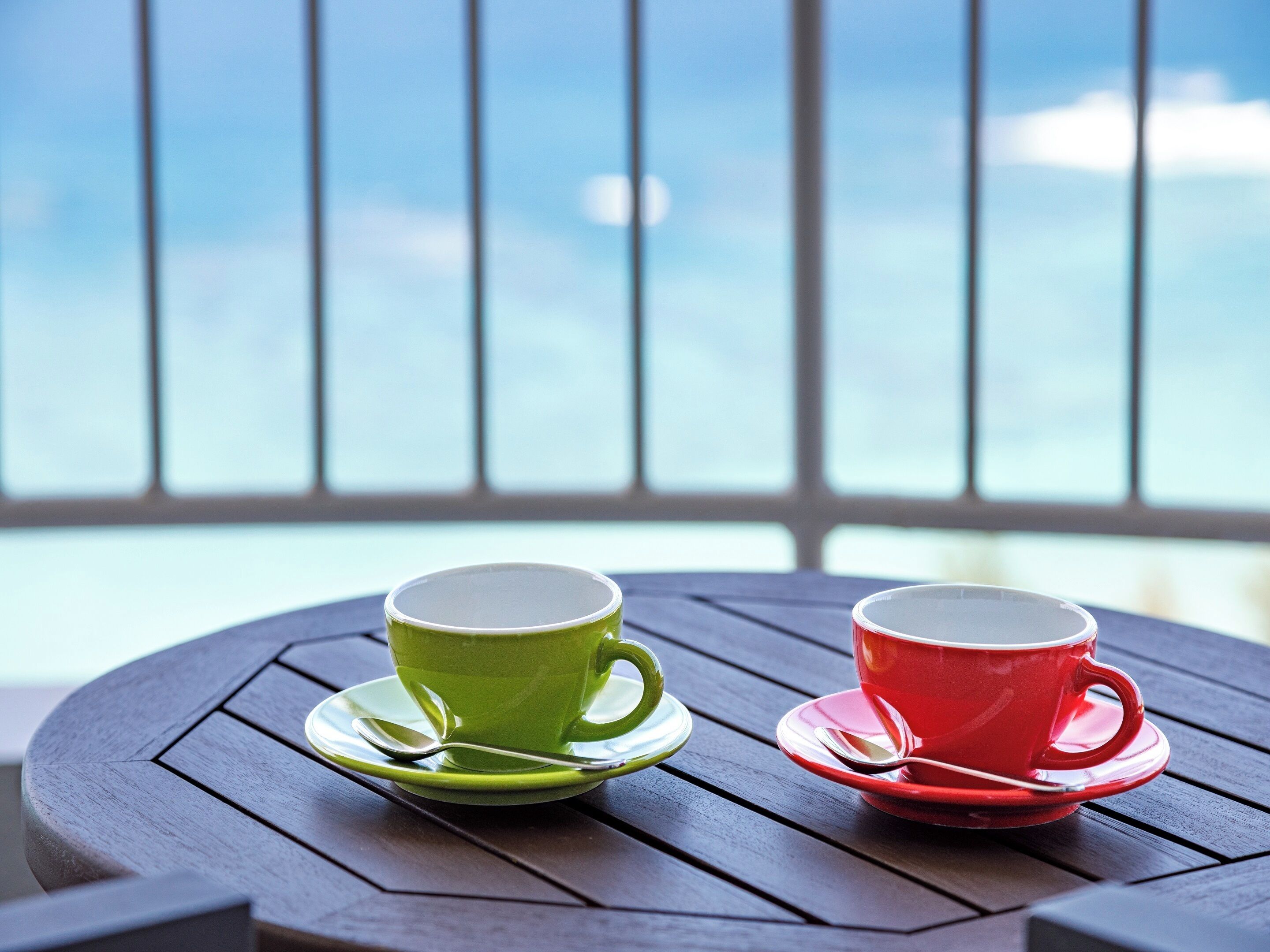 Two cups on a round wooden table with a view of the sky through a window.