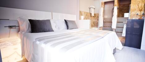 Una habitación de hotel moderna con una cama grande, un escritorio empotrado y un baño separado con pared de mármol y cabina de ducha de vidrio.