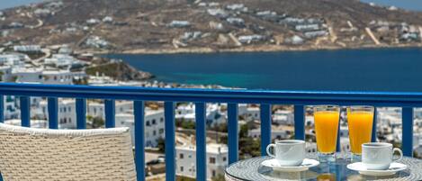A balcony with wicker chairs, a table with two glasses of orange juice, and a view of a coastal town.