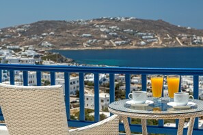 A balcony with wicker chairs, a table with two glasses of orange juice, and a view of a coastal town. - Hotel Alkyon (Mykonos)
