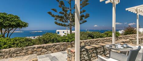 A terrace with stone walls, a pergola, wicker furniture, and a view of the sea and buildings.