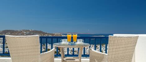 A balcony with wicker chairs and a table, overlooking a coastal town and the sea.