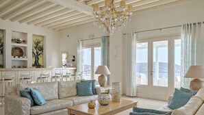 Amplia sala de estar con sofá, una mesa de centro y elementos decorativos. Se aprecia una vista al exterior a través de amplias ventanas.