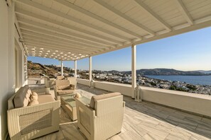 A covered outdoor seating area with wicker furniture and a view of a coastal town. - Hotel Alkyon (Mykonos)