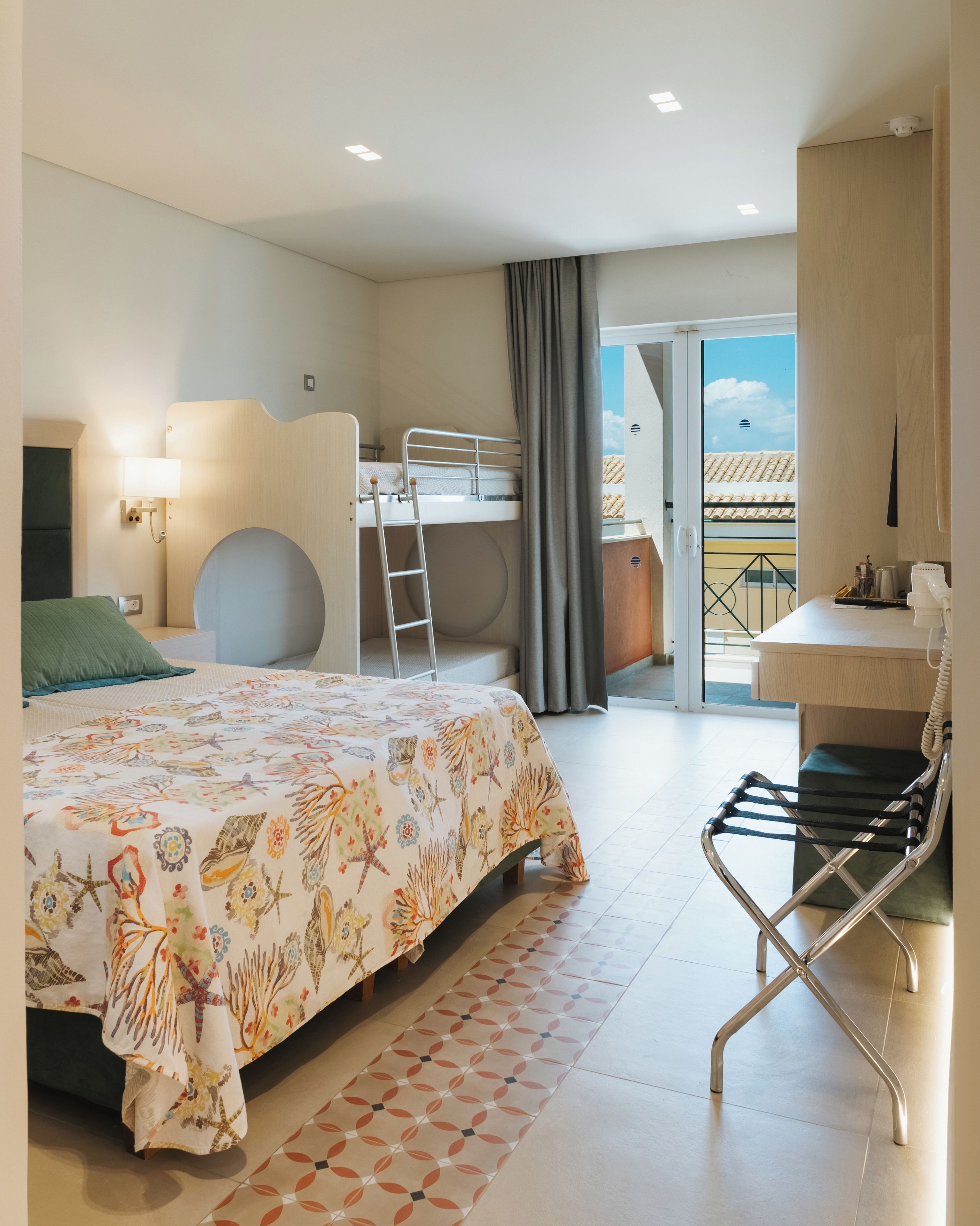 A hotel room with a bed, a desk, a chair, a bunk bed, and a balcony with a view.
