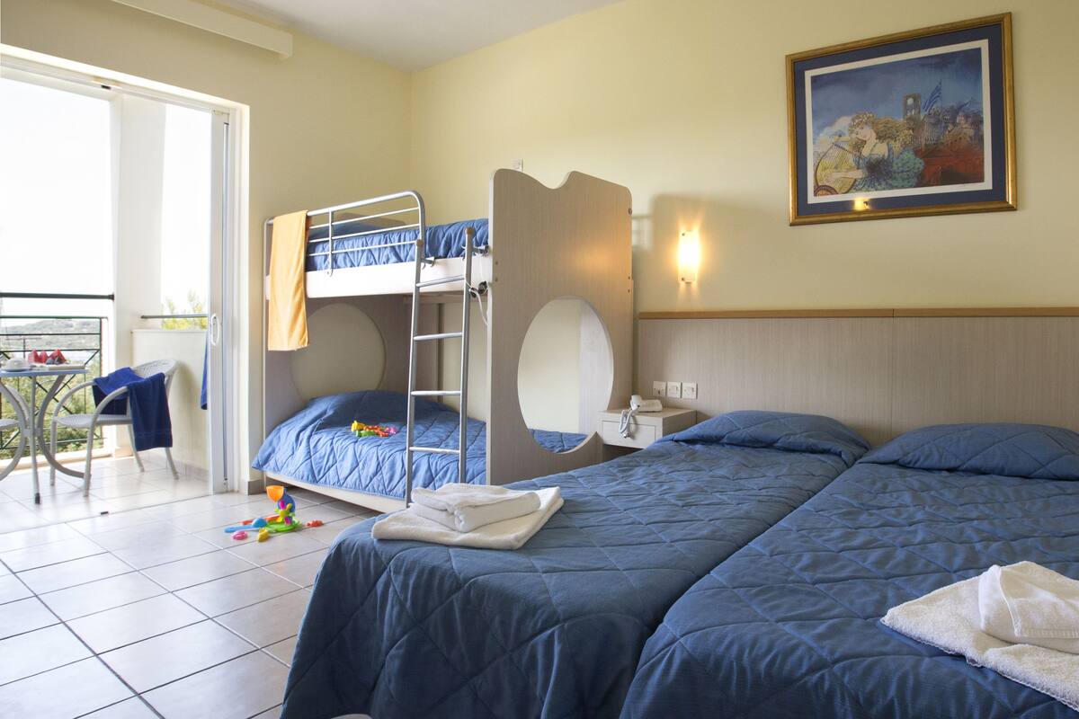 A hotel room with a bunk bed, a single bed, a small table with a chair, a bedside table with a lamp, and a painting on the wall.