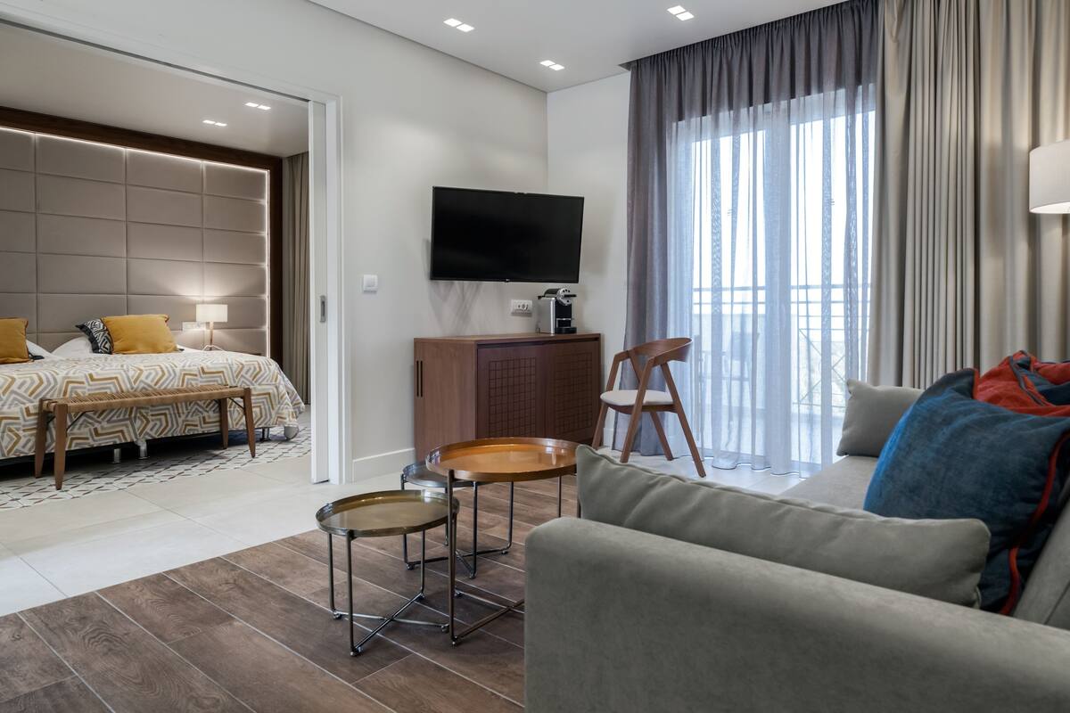 A modern hotel room with a bed, a sofa, a TV, a coffee machine, and a view of the city.