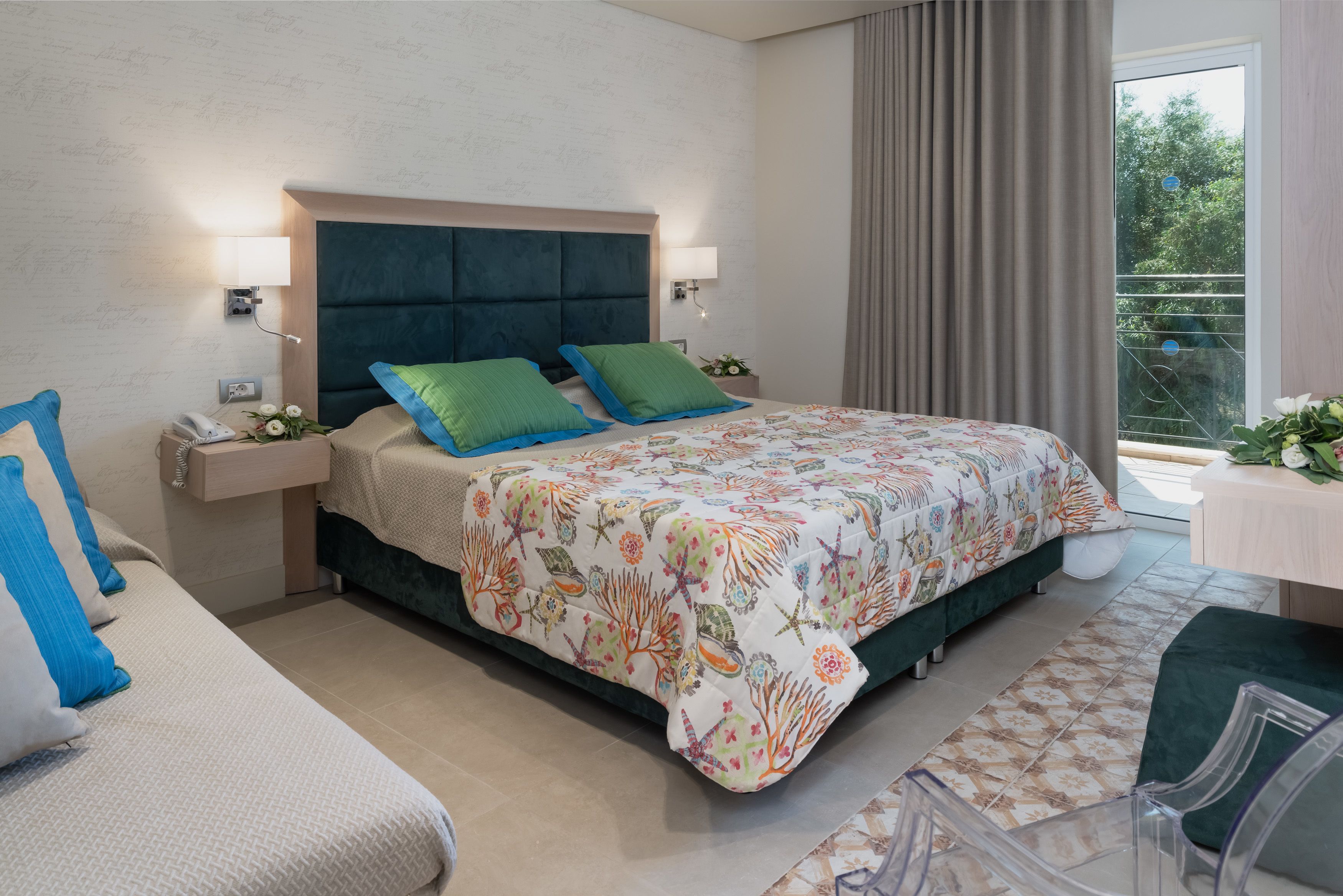 A modern hotel room with a large bed, a bedside table with a phone and flowers, a chair, and a view of greenery through the window.