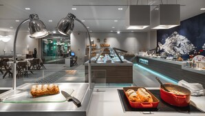 A modern buffet setup with a variety of food items, including a large piece of bread and a red dish, in a well-lit dining area with tables and chairs.