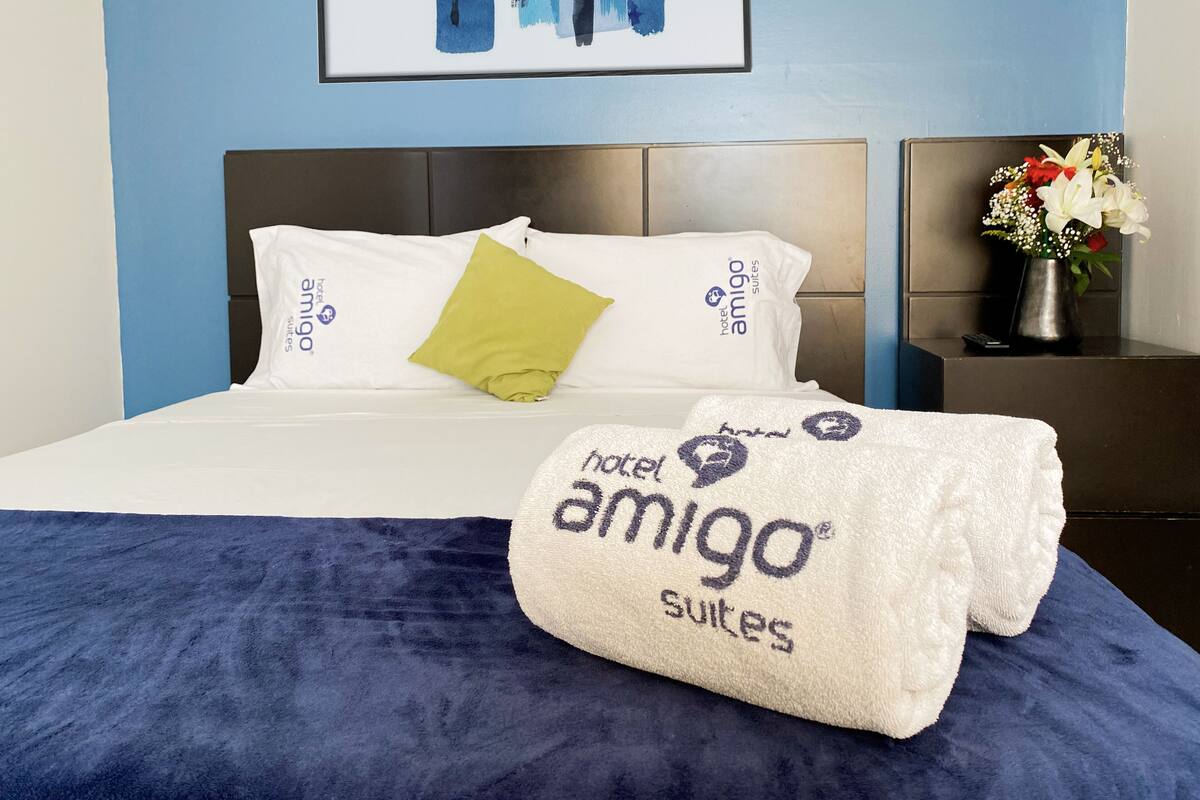 Habitación de hotel con cama, almohadas, manta, mesita de noche y un jarrón con flores.