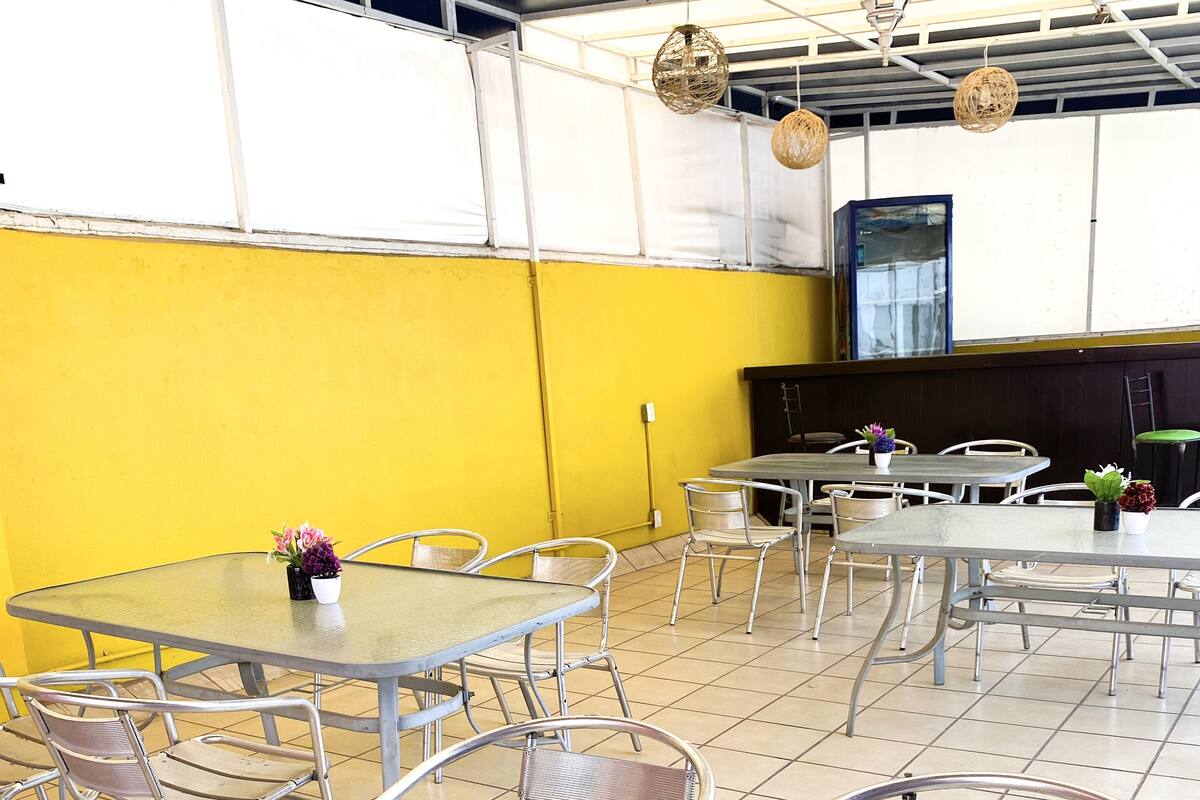 Un espacio exterior para comer con mesas y sillas de metal, rodeado de paredes amarillas y un suelo de baldosas.