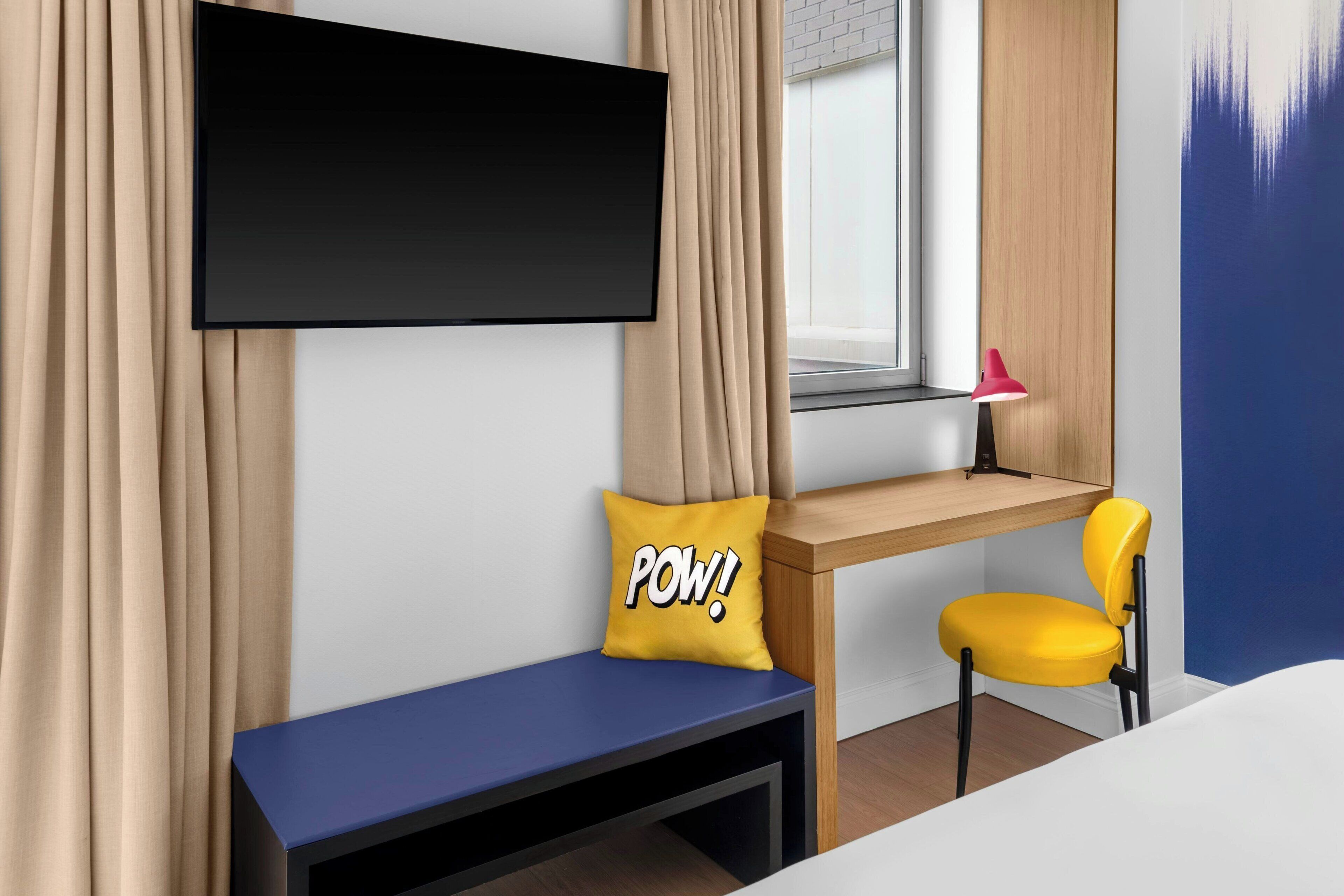 Une chambre d’hôtel moderne, équipée d’un lit, d’un bureau avec une chaise, d’un téléviseur fixé au mur et d’une fenêtre avec des rideaux.