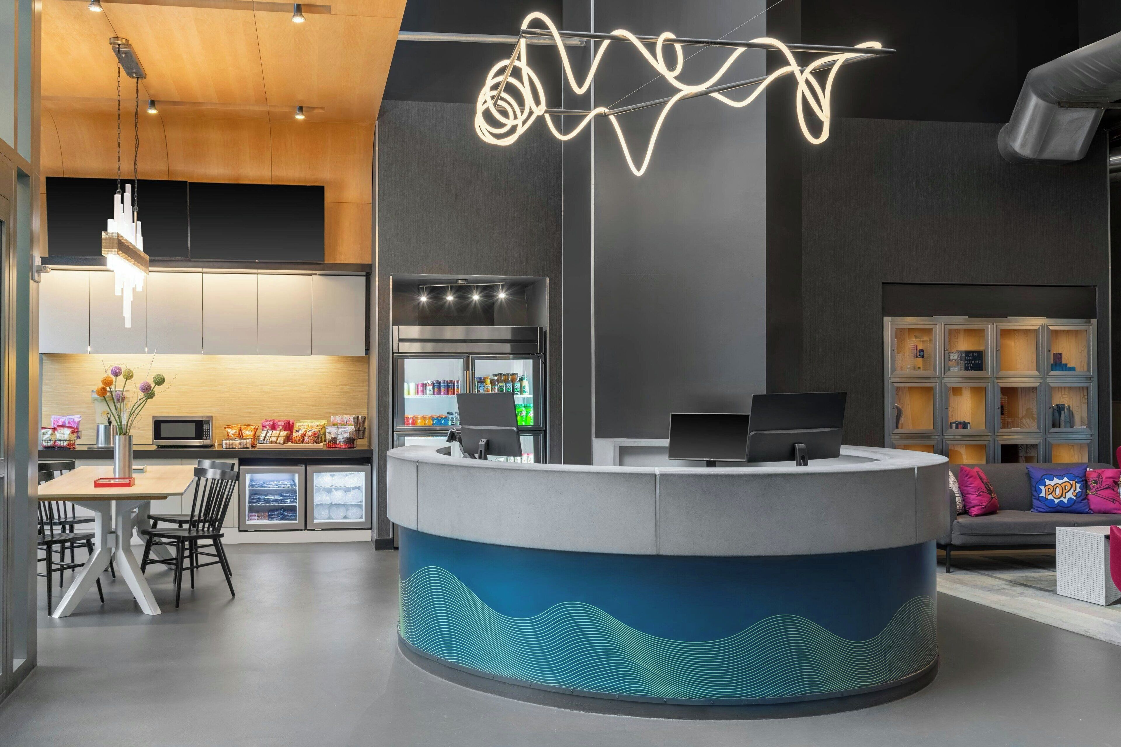 Uma área de recepção moderna com balcão curvo, sala de jantar com mesa e cadeiras, cozinha equipada com eletrodomésticos e geladeira, e um espaço de estar com sofá e estantes.