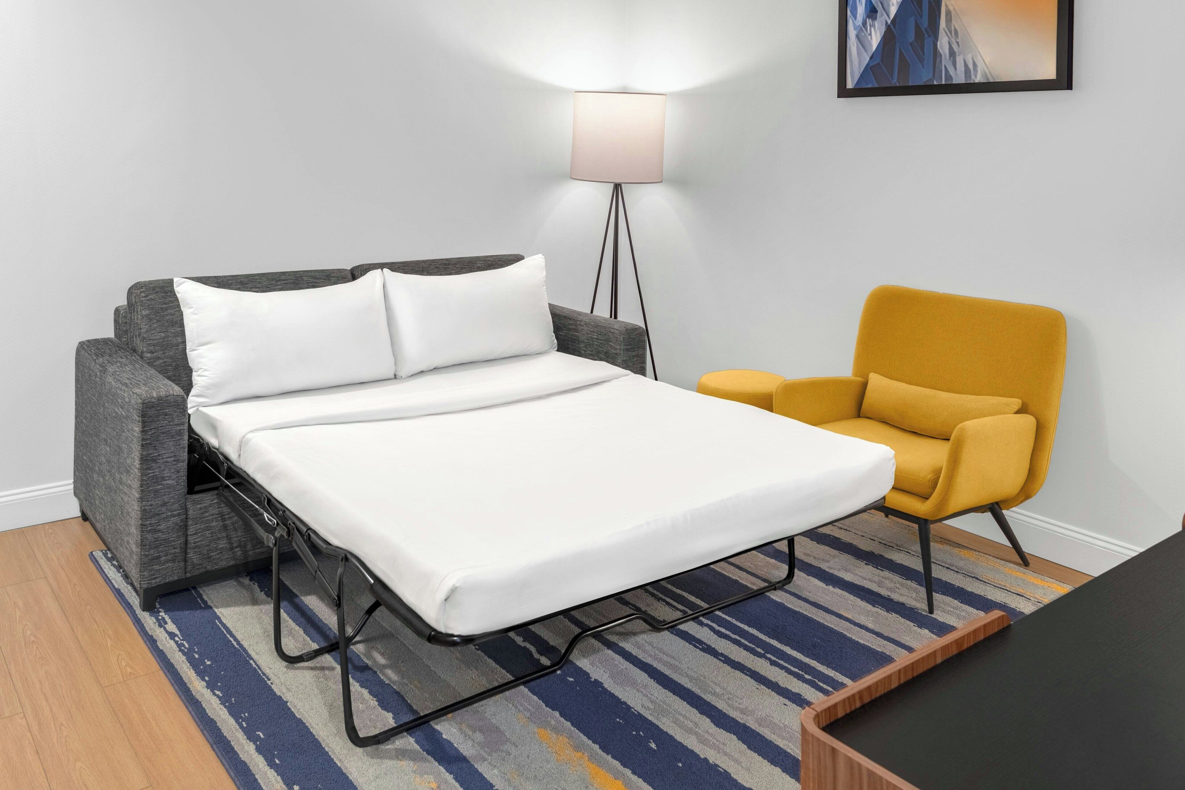 A compact bed with a white mattress and grey headboard, a mustard yellow armchair, a floor lamp, and a framed picture on the wall.