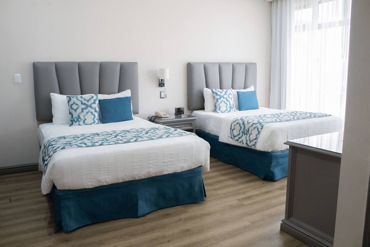 Ein Hotelzimmer mit zwei Betten, jeweils mit grauen Kopfteilen und blauer Bettwäsche. An den Betten stehen Nachttische mit Lampen und Telefonen. Der Raum hat Holzböden und ein Fenster mit durchsichtigen Vorhängen.