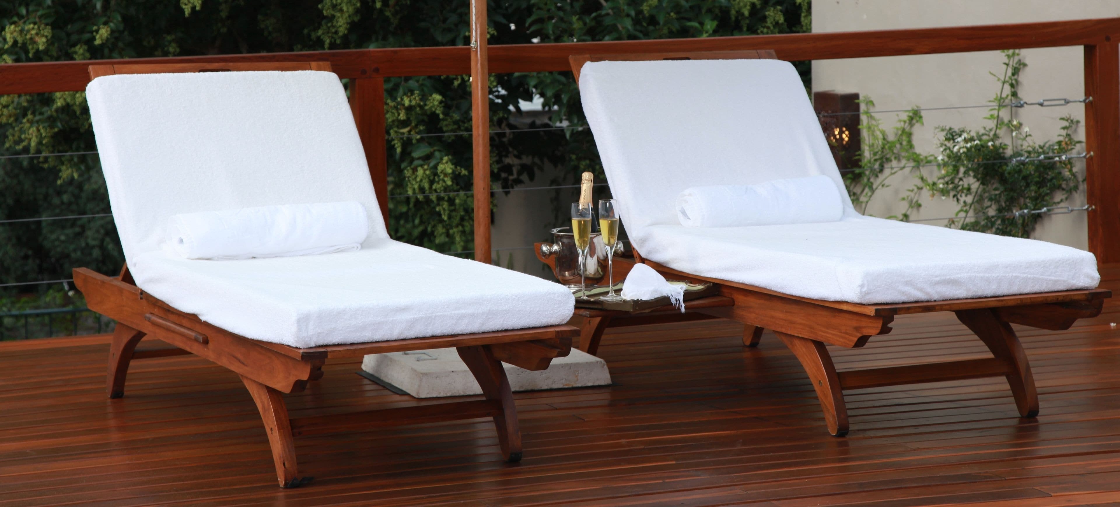 Two wooden lounge chairs with white cushions and a small table with a bottle and glasses.