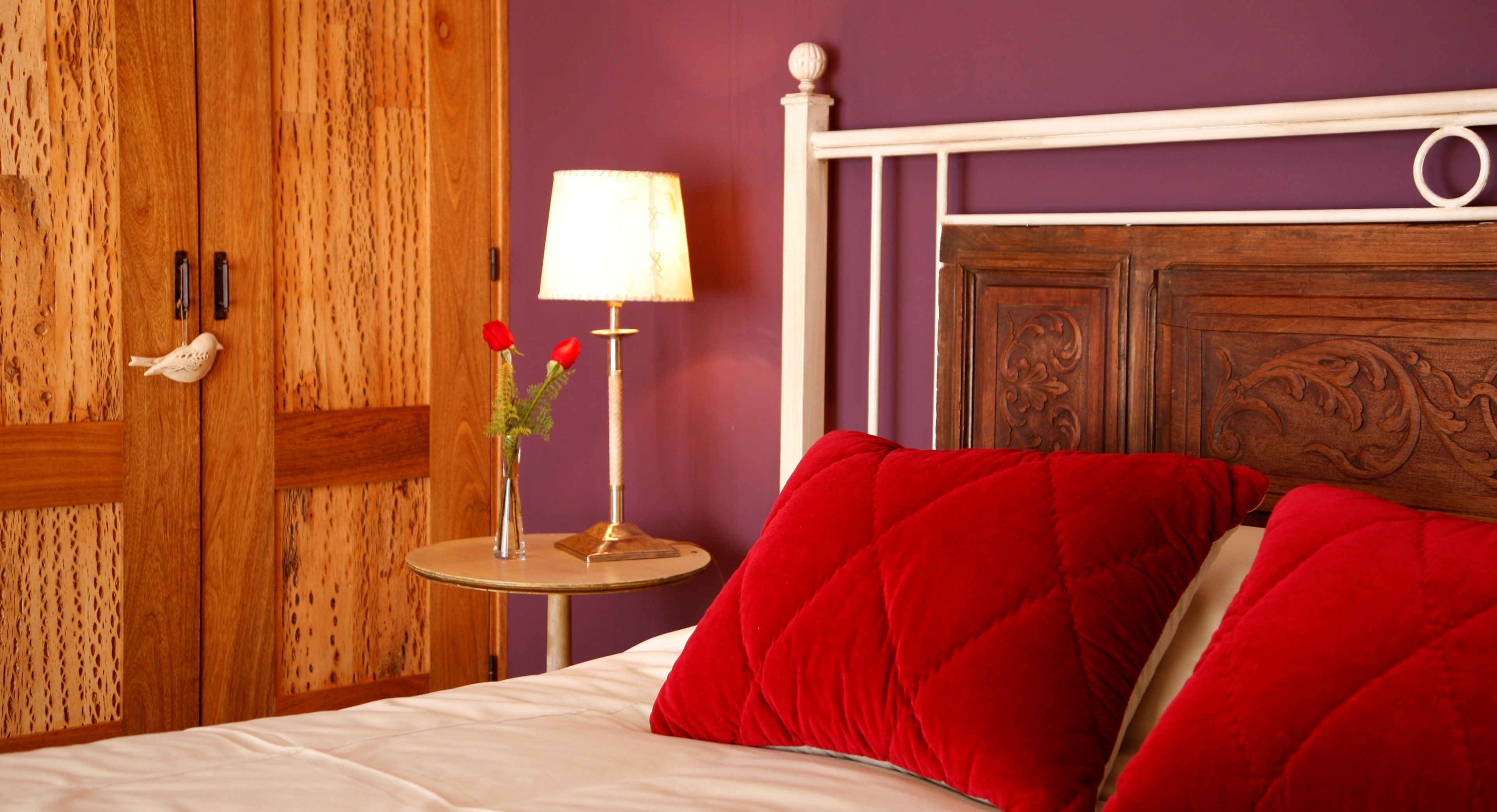 A bedroom with a wooden wardrobe, a bedside table with a lamp, and red pillows.