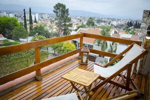 Una terraza de madera con una mesa y dos sillones de descanso, con vistas a un paisaje urbano de edificios y árboles.