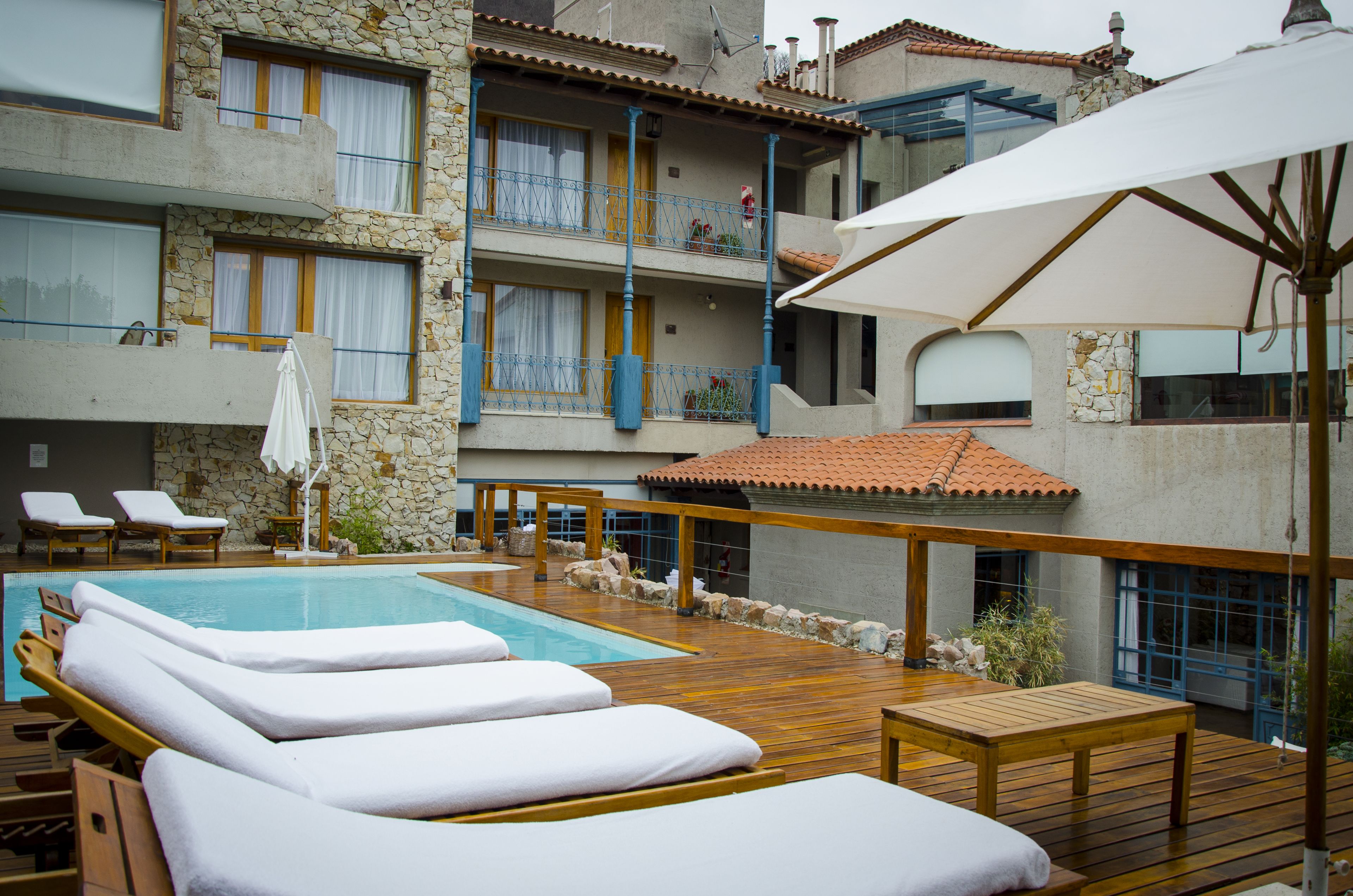 Un área junto a la piscina con sillones de descanso, una terraza de madera y un edificio de piedra con balcones.
