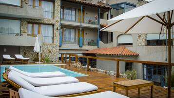 Un área junto a la piscina con sillones de descanso, una terraza de madera y un edificio de piedra con balcones.