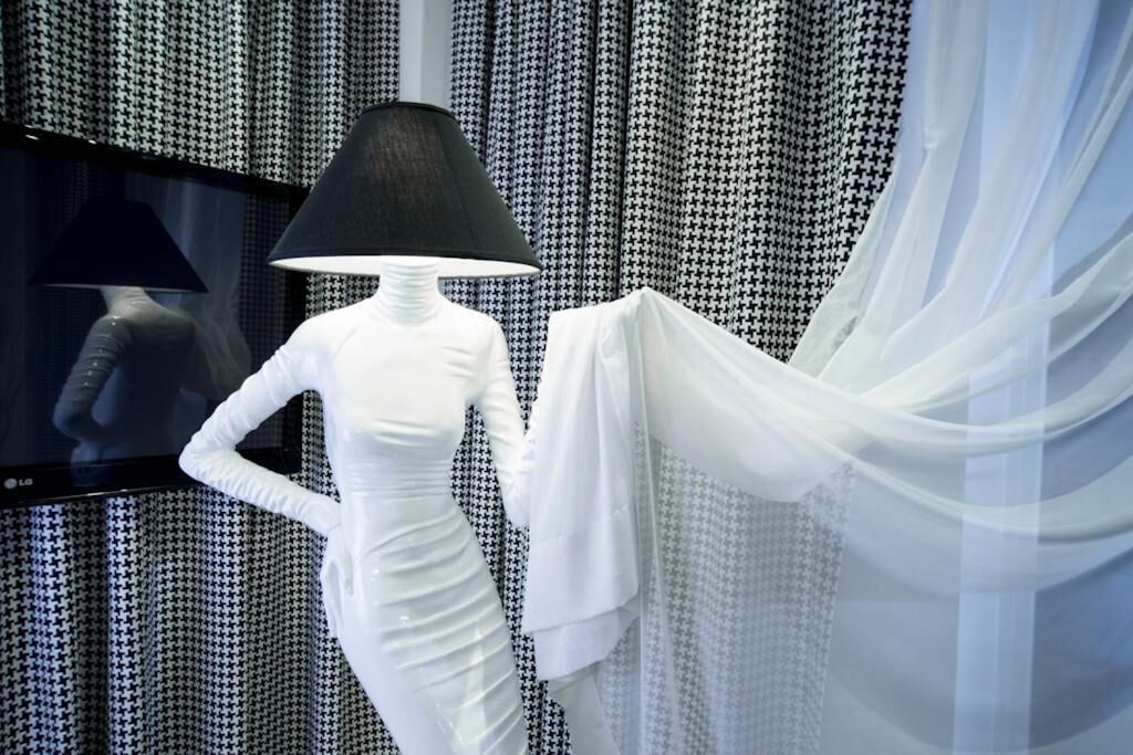 A mannequin in a white dress with a black lampshade, positioned in front of a patterned backdrop.