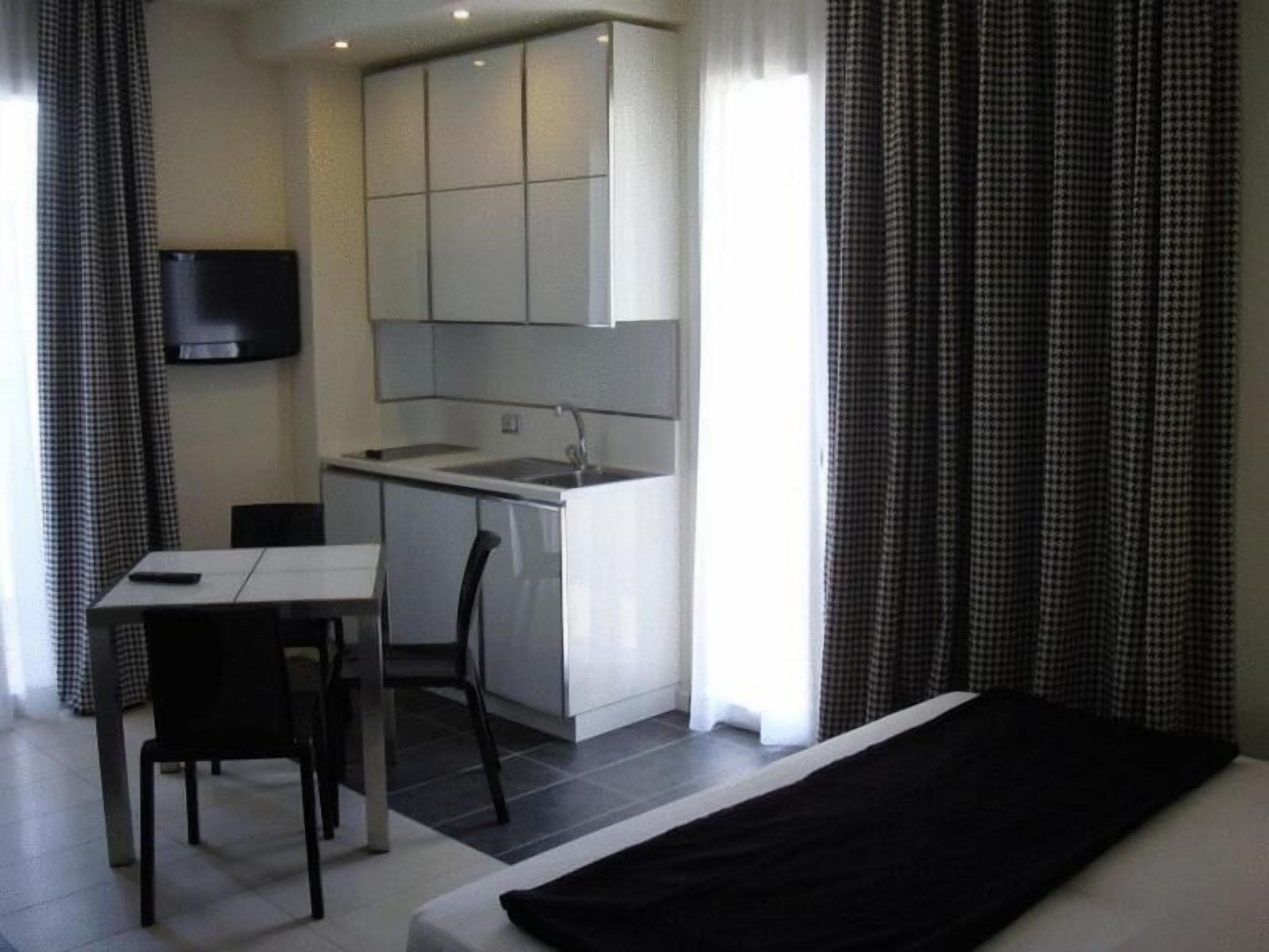 A compact hotel room with a kitchenette, a single bed, a small table with a chair, and a TV mounted on the wall.