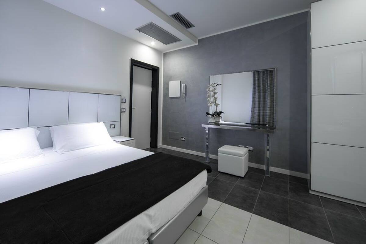 Une chambre moderne avec un grand lit, une table de chevet, une commode, une petite table d’appoint, un miroir, une étagère murale agrémentée d’une plante et une porte.