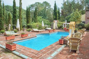 Une piscine rectangulaire dont le bord est recouvert de carreaux, entourée d’un sol en briques et de plantes en pot.
