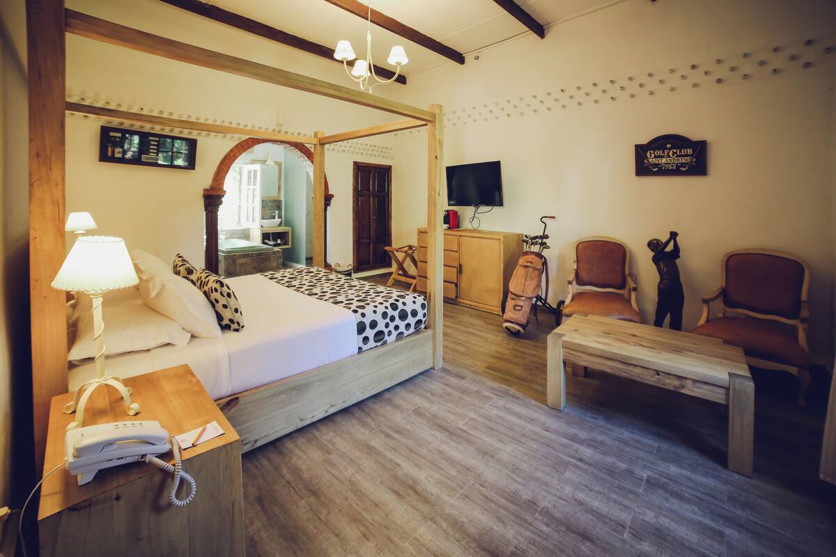 Habitación de hotel con una cama, una mesita de noche de madera con teléfono, un escritorio con lámpara, televisión y un cartel con el logo de un club de golf en la pared.