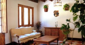 Un salon chaleureux avec un canapé, une table en bois et des meubles de rangement. Des plantes en pot sont disposées sur le mur, et une fenêtre est équipée de stores.