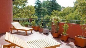 Un balcón con dos sillones de madera, varias plantas en macetas y vistas a árboles y edificios.