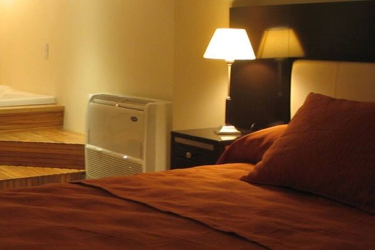 A hotel room with a wooden bath tub, a bed with a red blanket, a nightstand with a lamp, and a wall-mounted heater.