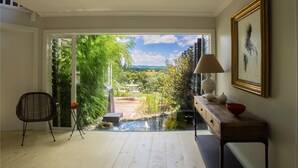 Una habitación con piso de madera, una silla, una mesa con lámpara, un cuadro enmarcado y vista a un jardín con estanque.