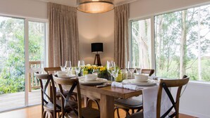 Comedor con mesa de madera dispuesta para cuatro personas, sillas, una lámpara colgante y vistas a la vegetación a través de amplios ventanales.