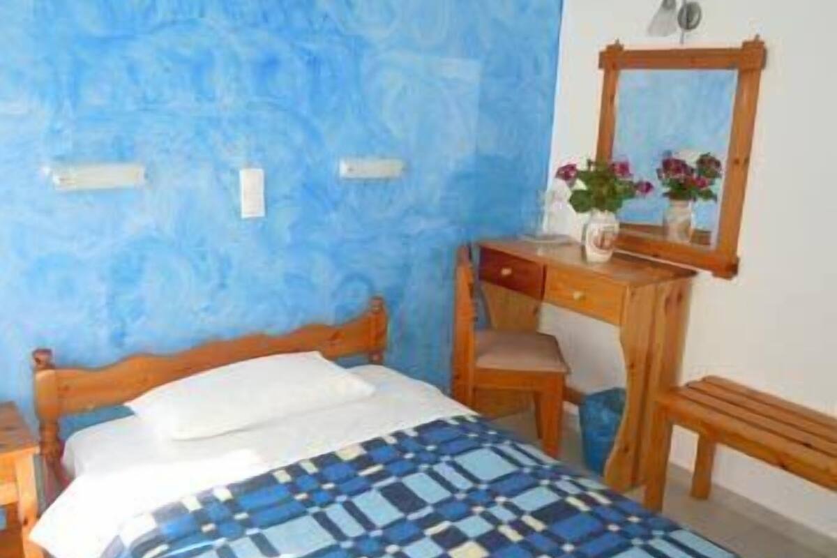 Un dormitorio con pared azul con textura, estructura de cama de madera, colcha estampada, mesita de noche de madera, cómoda de madera, espejo y una pequeña mesa con un jarrón de flores.
