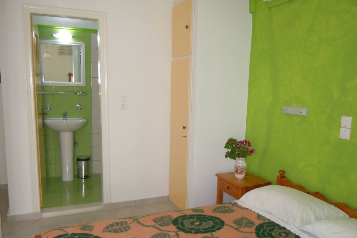 Un dormitorio con cama, una mesita de noche con un florero, una pared verde y un baño visible a través de una puerta abierta.