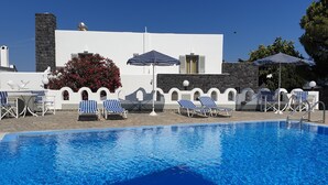 Piscina com espreguiçadeiras e guarda-sóis, cercada por um edifício branco com detalhes em pedra e um céu azul límpido.