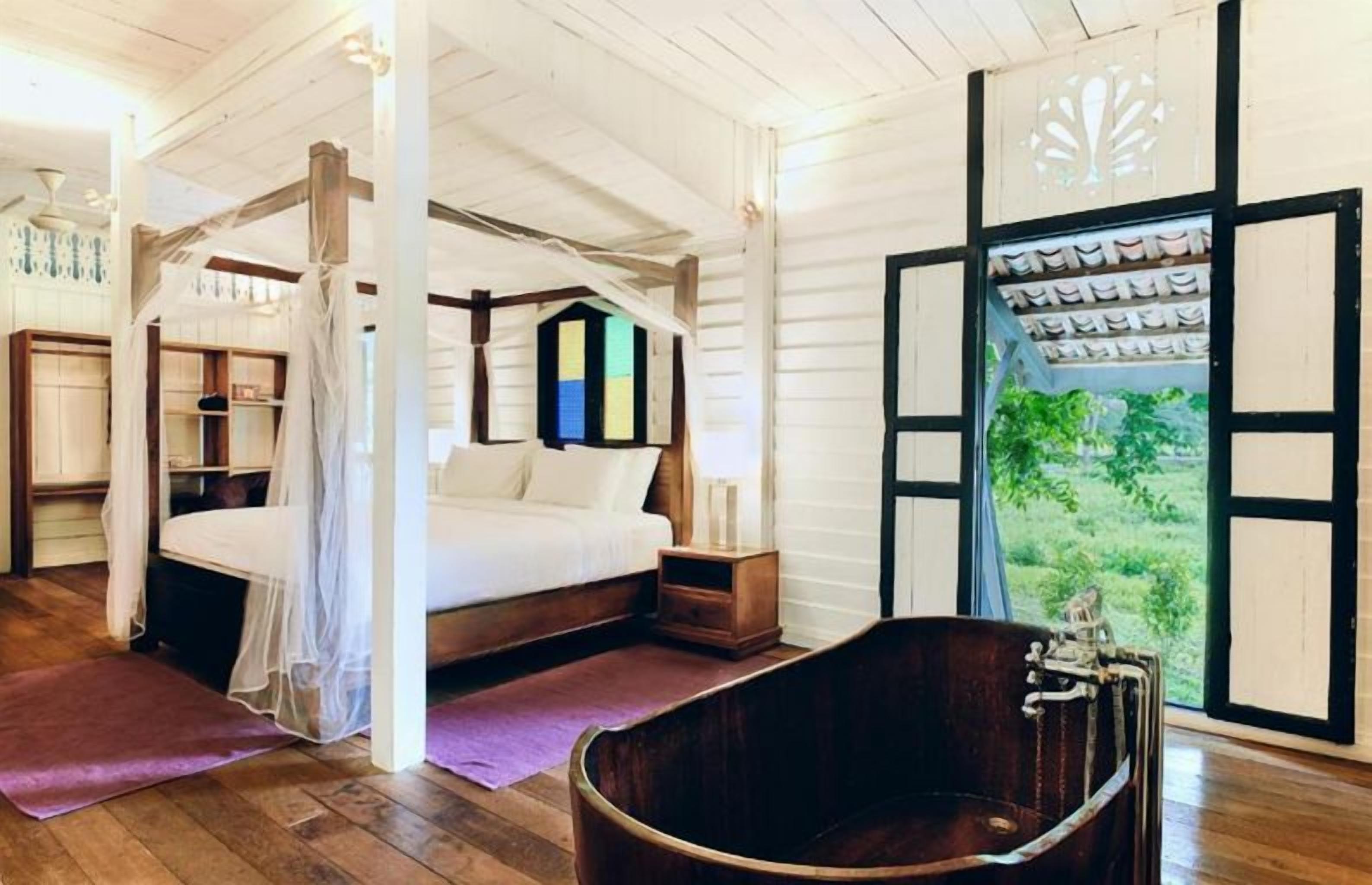 A bedroom with a canopy bed, a wooden bathtub, and a view of greenery outside.