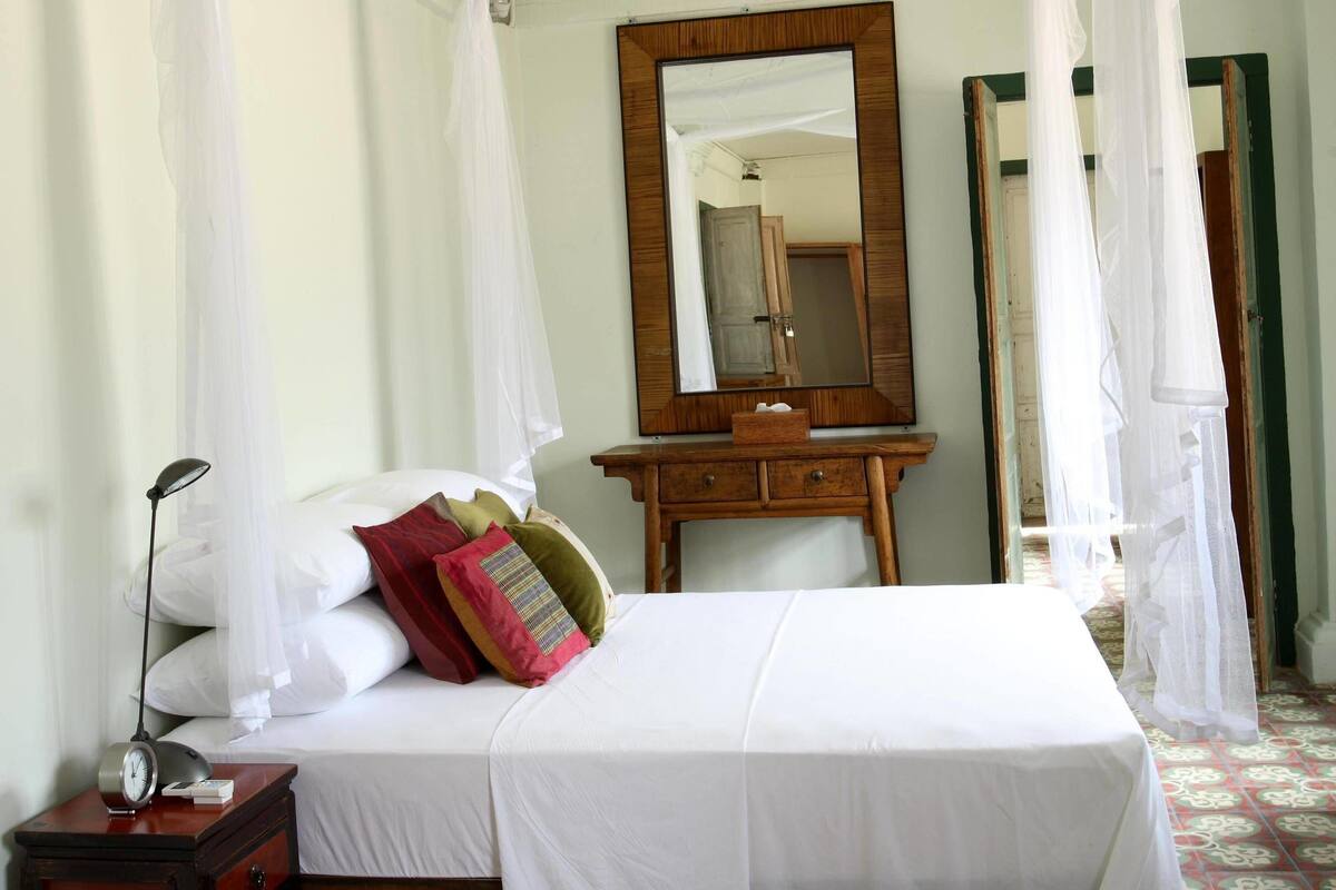 A bedroom with a four-poster bed, a wooden dresser, a mirror, and a bedside table with a lamp.