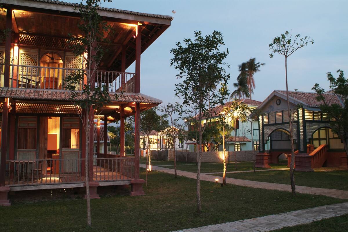 A traditional wooden building with a balcony, surrounded by a well-maintained garden and palm trees.